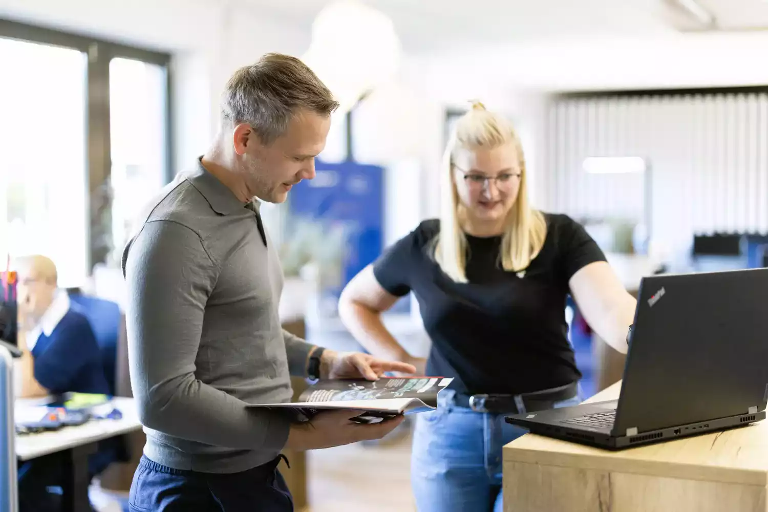 Mitarbeiter besprechen Projekt im Büro am Laptop, Kunde hält Broschüre in Hand